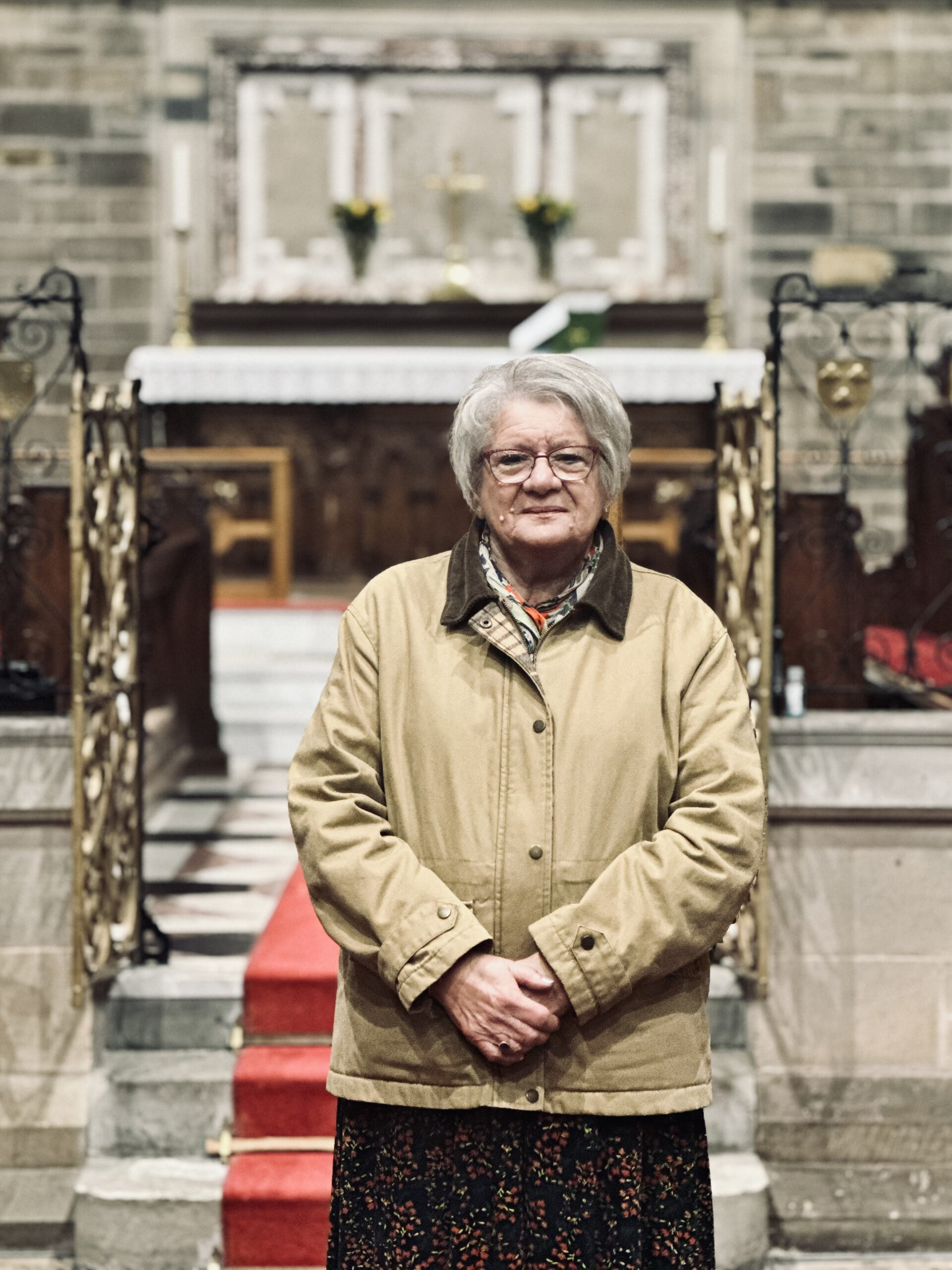 An elderly woman with grey hair and glasses, wearing a beige coat and patterned skirt, stands on a red carpet in front of an ornate church altar.