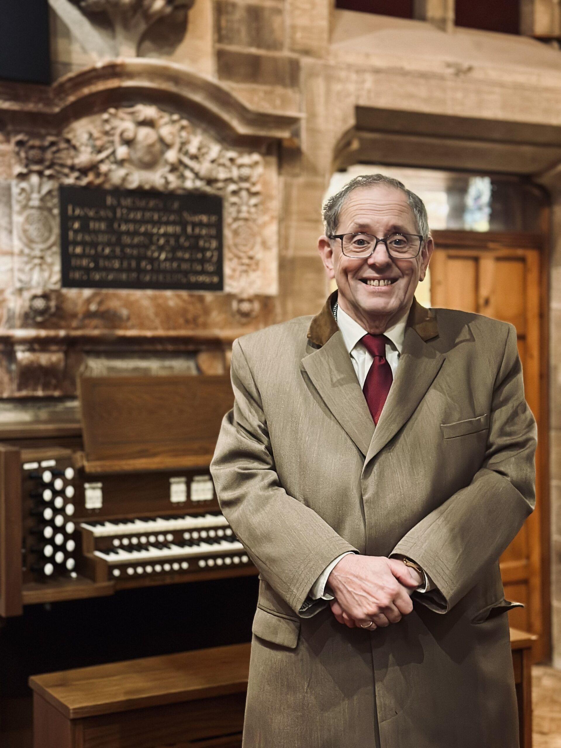 A smiling man in a tan suit and red tie stands in front of a large pipe organ and a stone memorial plaque inside a church with stone walls.
