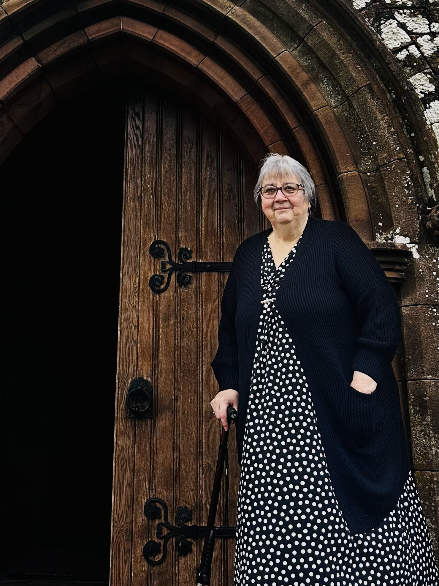 An older woman with grey hair, glasses, and a walking stick stands in front of a large wooden arched door. She wears a black cardigan over a black and white polka dot dress and smiles slightly.