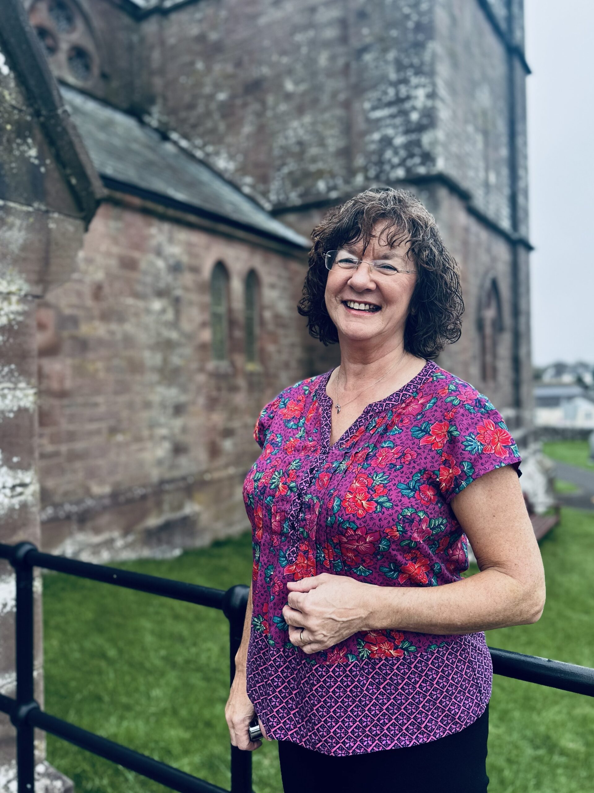 A smiling woman with curly hair, wearing glasses and a colourful floral blouse, stands outdoors in front of an old stone building with arched windows and green grass.