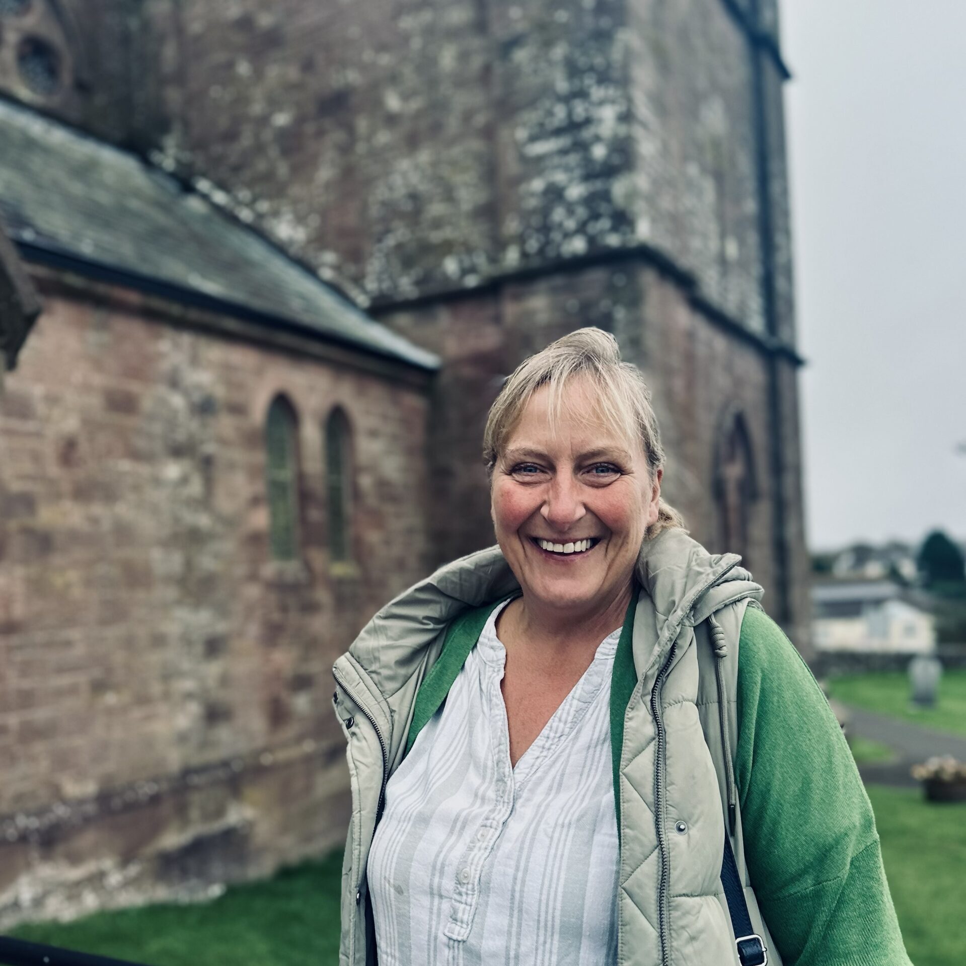 A smiling woman in a light green jacket and white shirt stands outside near an old stone building with arched windows and a grassy area on a cloudy day.