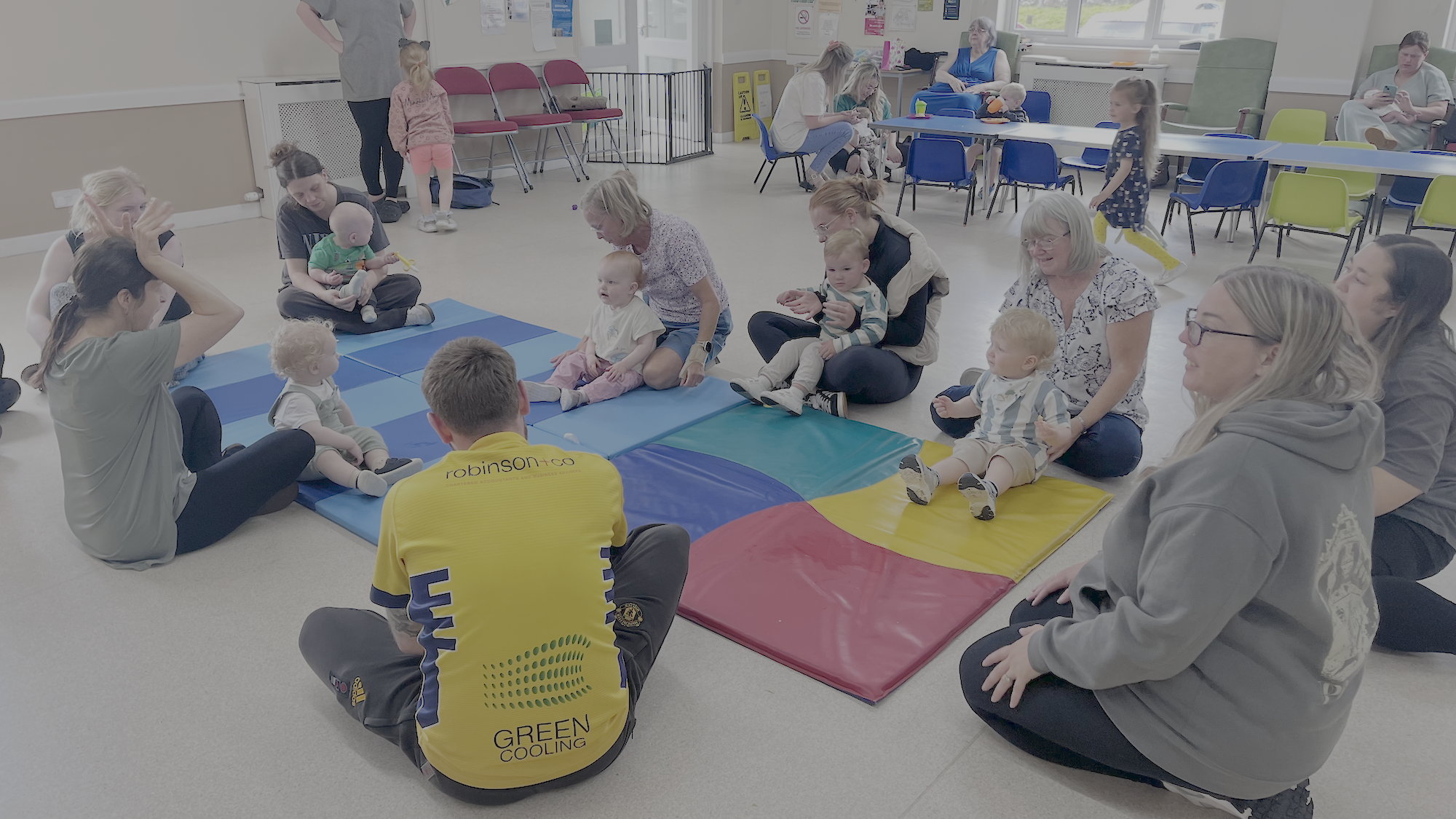 A group of adults and babies sit in a circle on colourful mats in a bright room, engaging in a group activity. More adults and children are seated at tables in the background.