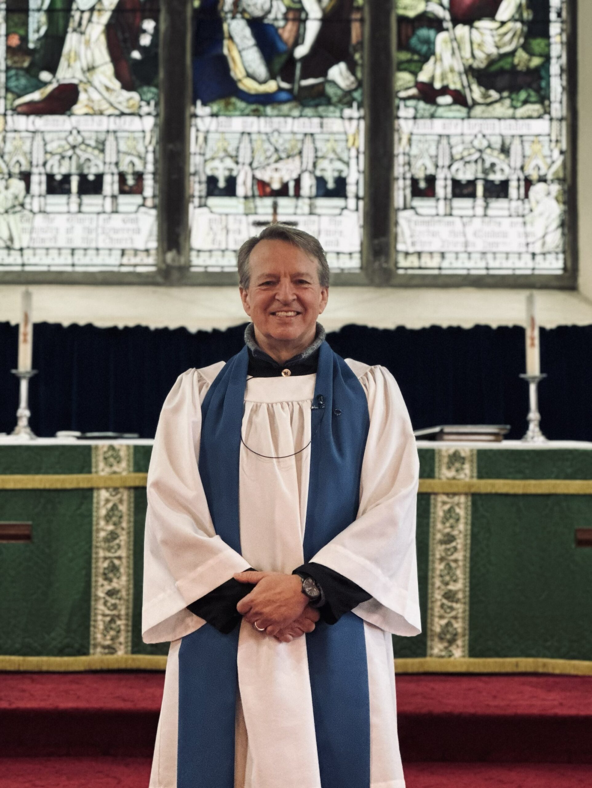 A smiling member of the clergy wearing white and blue robes stands in front of an altar with green cloth, candles, and stained glass windows in a church.