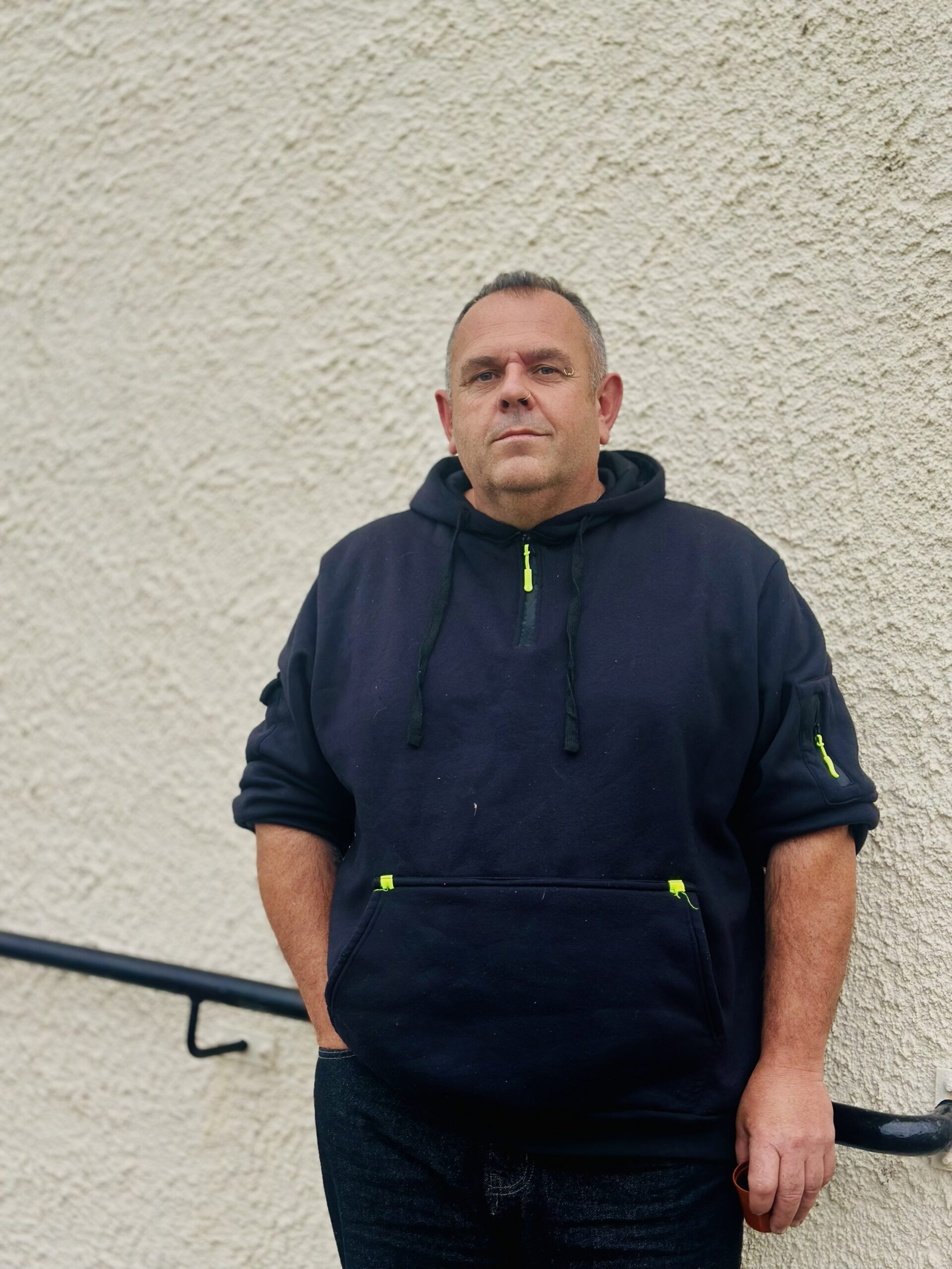 A man wearing a dark hoodie stands against a textured, light-coloured wall with one hand in his pocket, looking at the camera with a neutral expression. A black handrail runs along the wall beside him.