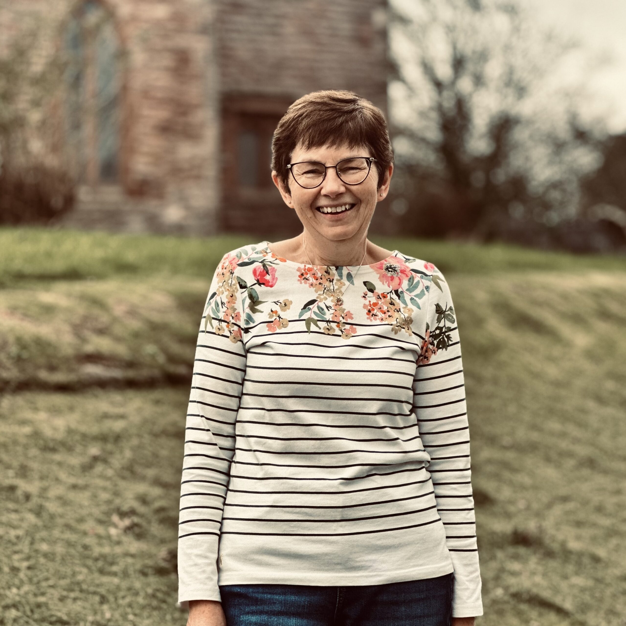 A woman with short brown hair and glasses smiles whilst standing outdoors on grass, wearing a striped and floral long-sleeved shirt. A blurred building and trees are visible in the background.