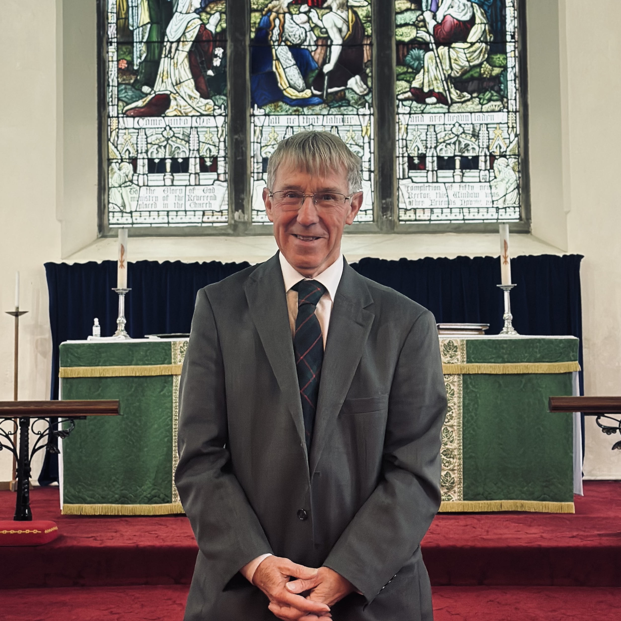 An older man in a suit and tie stands smiling in front of an altar with a green cloth in a church, with stained glass windows in the background.