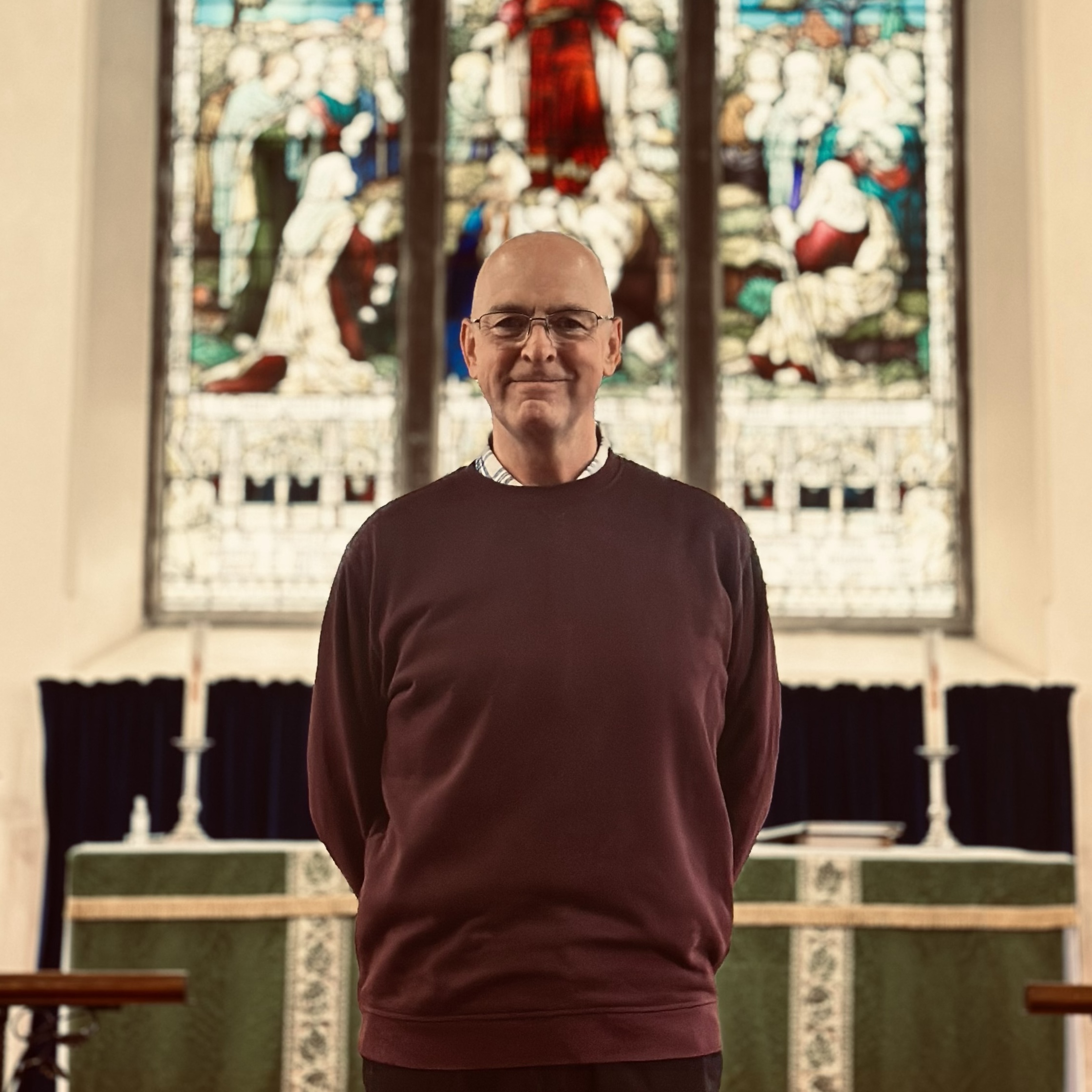 A man wearing glasses and a maroon jumper stands in front of an altar with green cloth and two candles, with stained glass windows in the background.