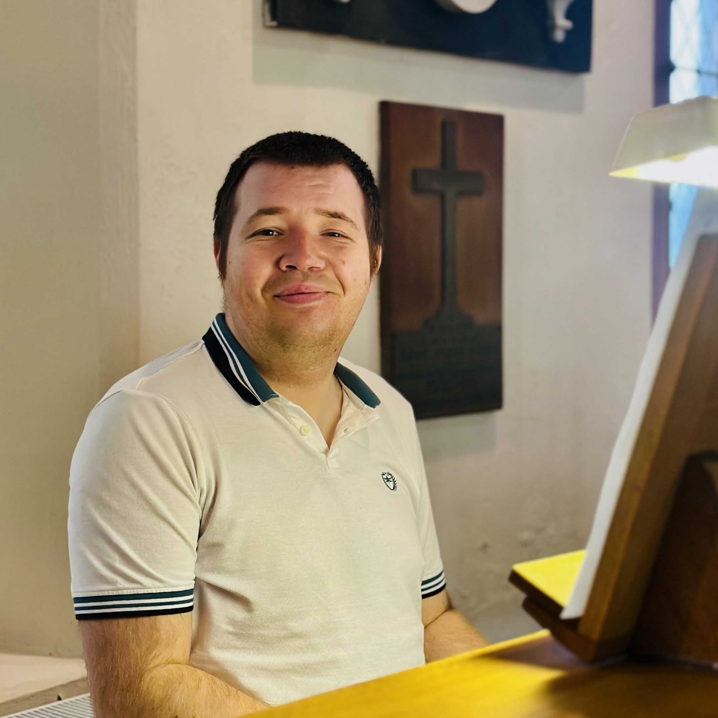 A man in a white polo shirt sits at a wooden piano or organ, smiling at the camera. Behind him is a wall plaque with a cross and a lit lamp, suggesting a church or similar setting.