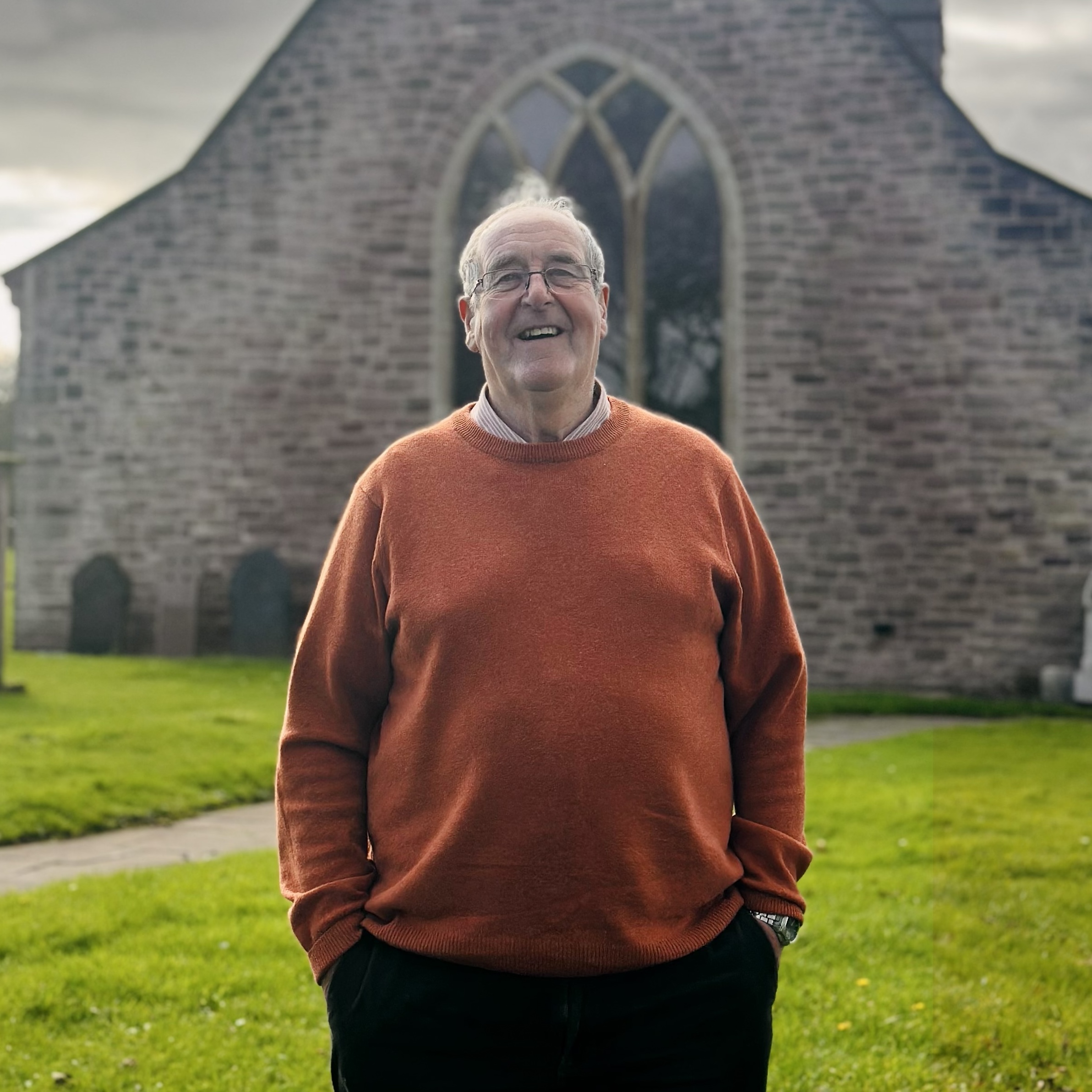 An older man wearing glasses and an orange jumper stands smiling outside in front of an old stone church with arched windows and a grassy garden.