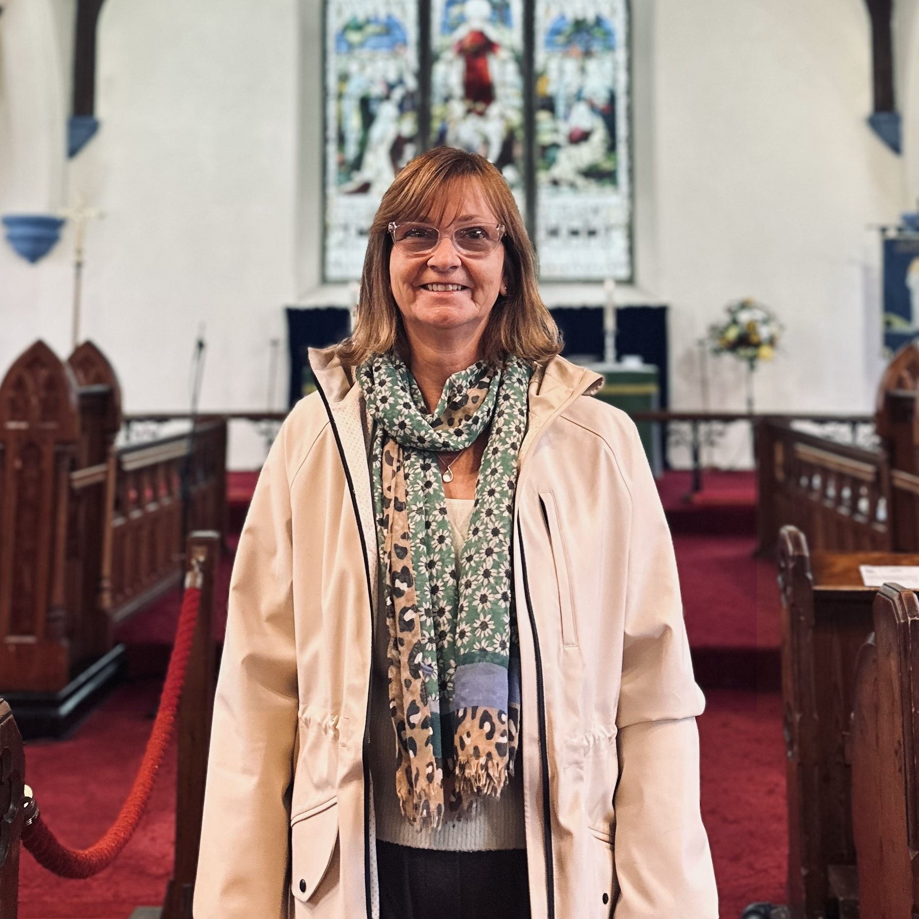 A woman wearing glasses, a light-coloured jacket, and a patterned scarf stands smiling inside a church with red carpet, wooden pews, and stained glass windows in the background.