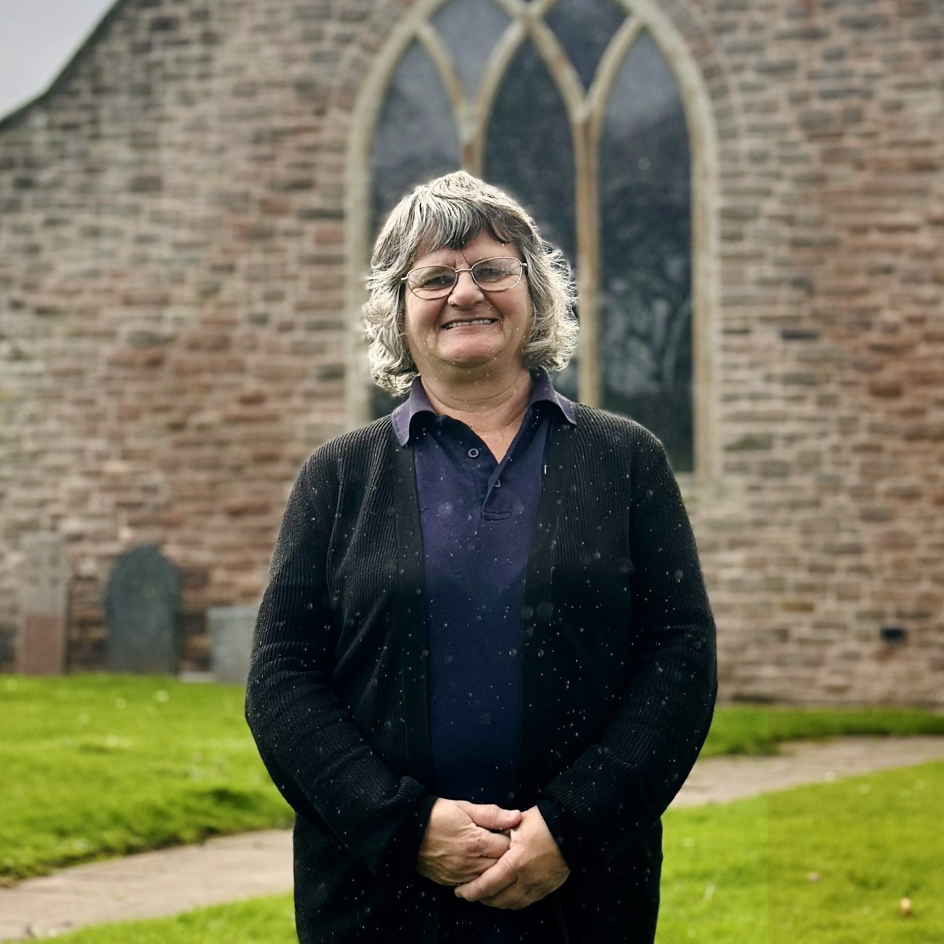 A smiling woman with glasses and short grey hair stands outside in front of a stone building with arched windows, on a grassy area with a stone path.