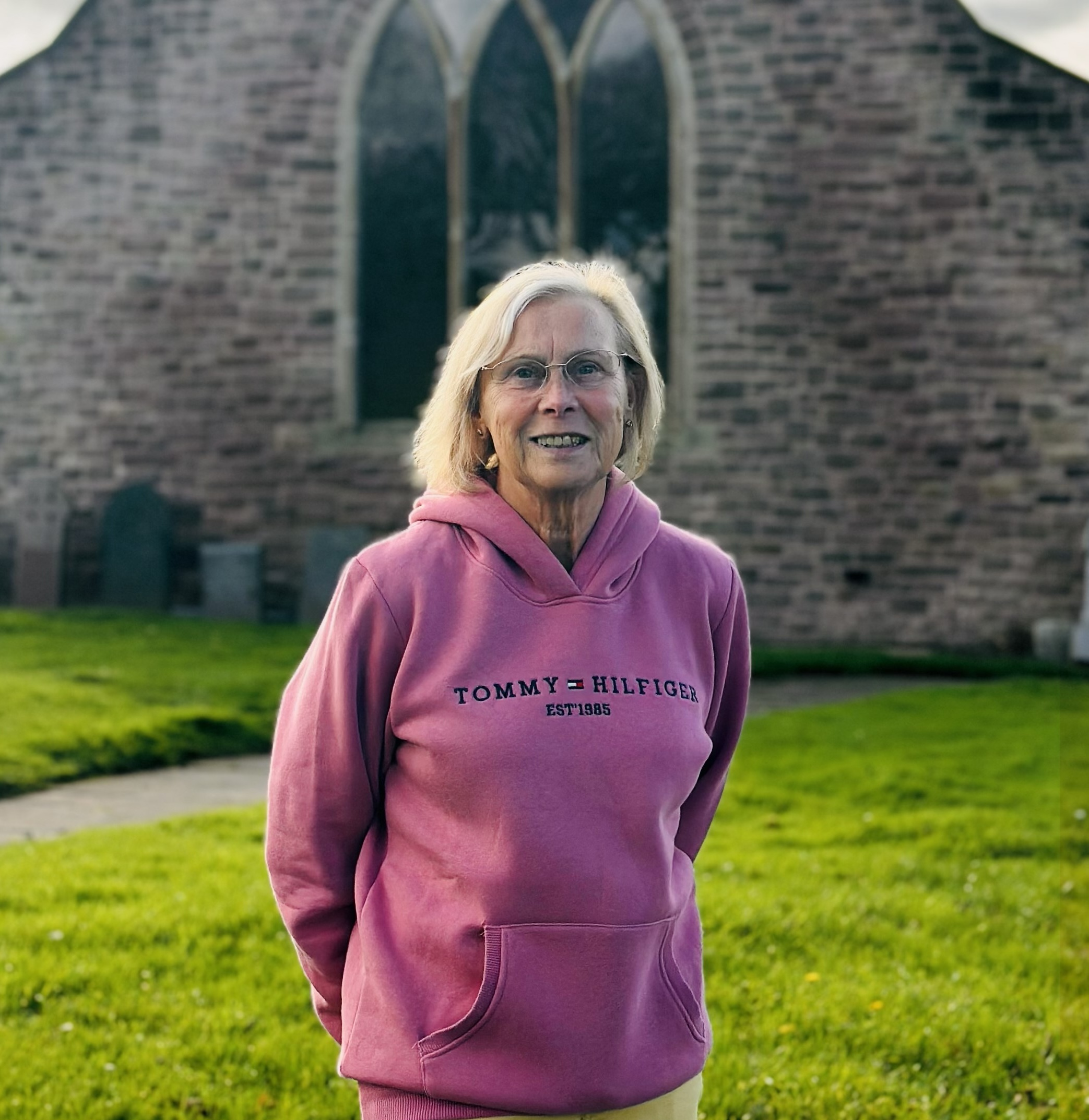 An older woman with glasses and a pink Tommy Hilfiger hoodie stands smiling outside in front of a stone building with arched windows and a green lawn.