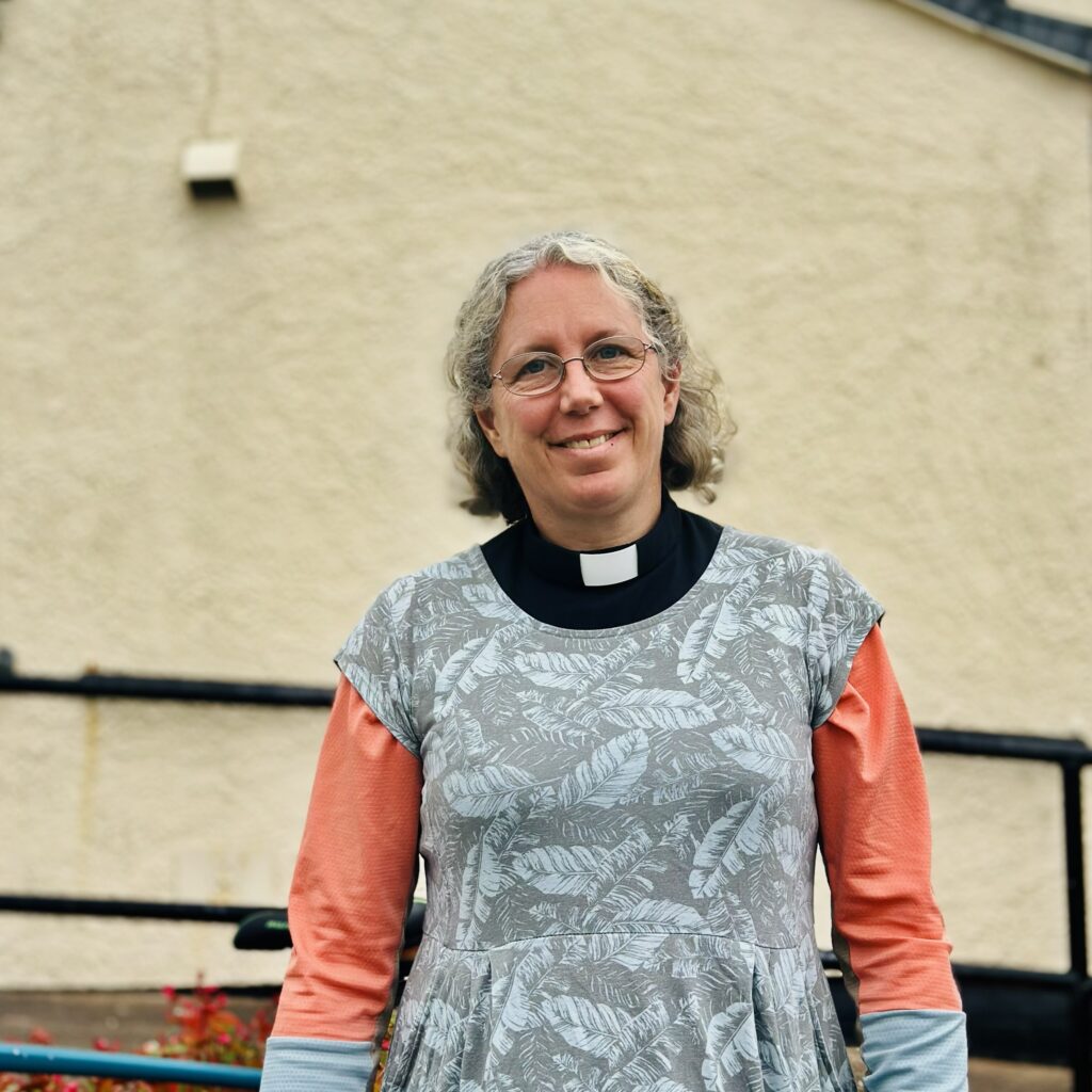 A smiling woman with grey curly hair and glasses, wearing a clerical collar, a grey leaf-patterned dress, and an orange long-sleeved shirt, stands in front of a beige textured wall and black railing.