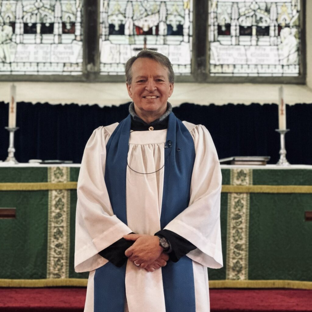 A smiling clergyman in white and blue robes stands in front of an altar with green cloths and tall candles, with stained glass windows in the background.