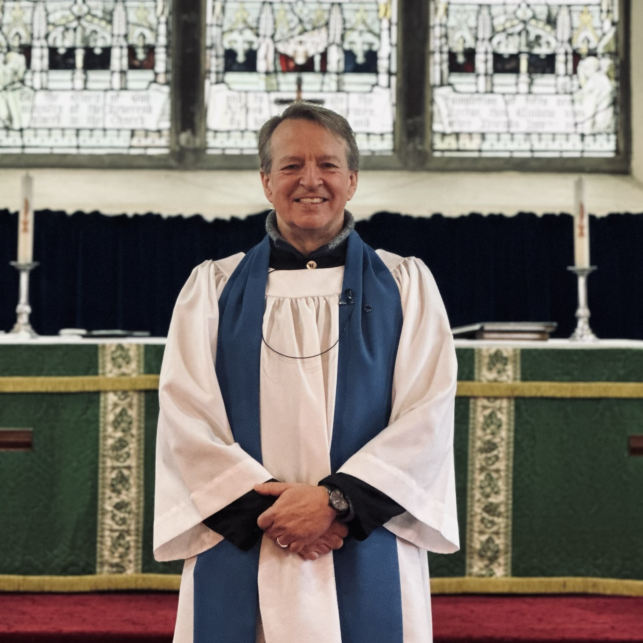 A smiling clergyman in white and blue robes stands in front of an altar with green cloths and tall candles, with stained glass windows in the background.