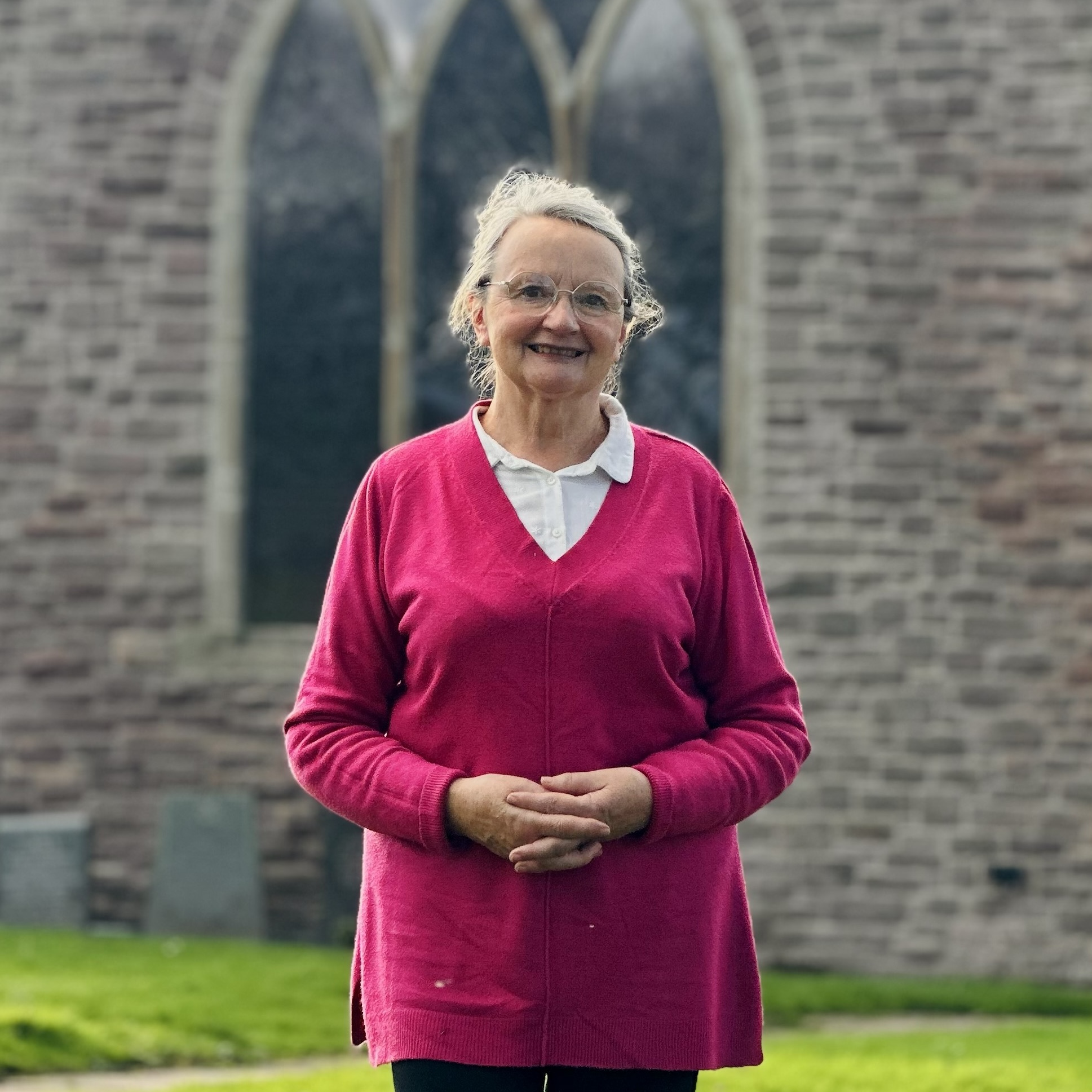 An older woman with glasses, grey hair, and a pink jumper stands smiling in front of a stone building with arched windows and grass in the background.