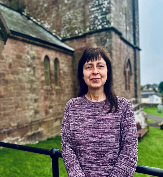 A woman with straight brown hair and a purple jumper stands outside in front of an old stone building with arched windows, grassy lawn, and cloudy sky.