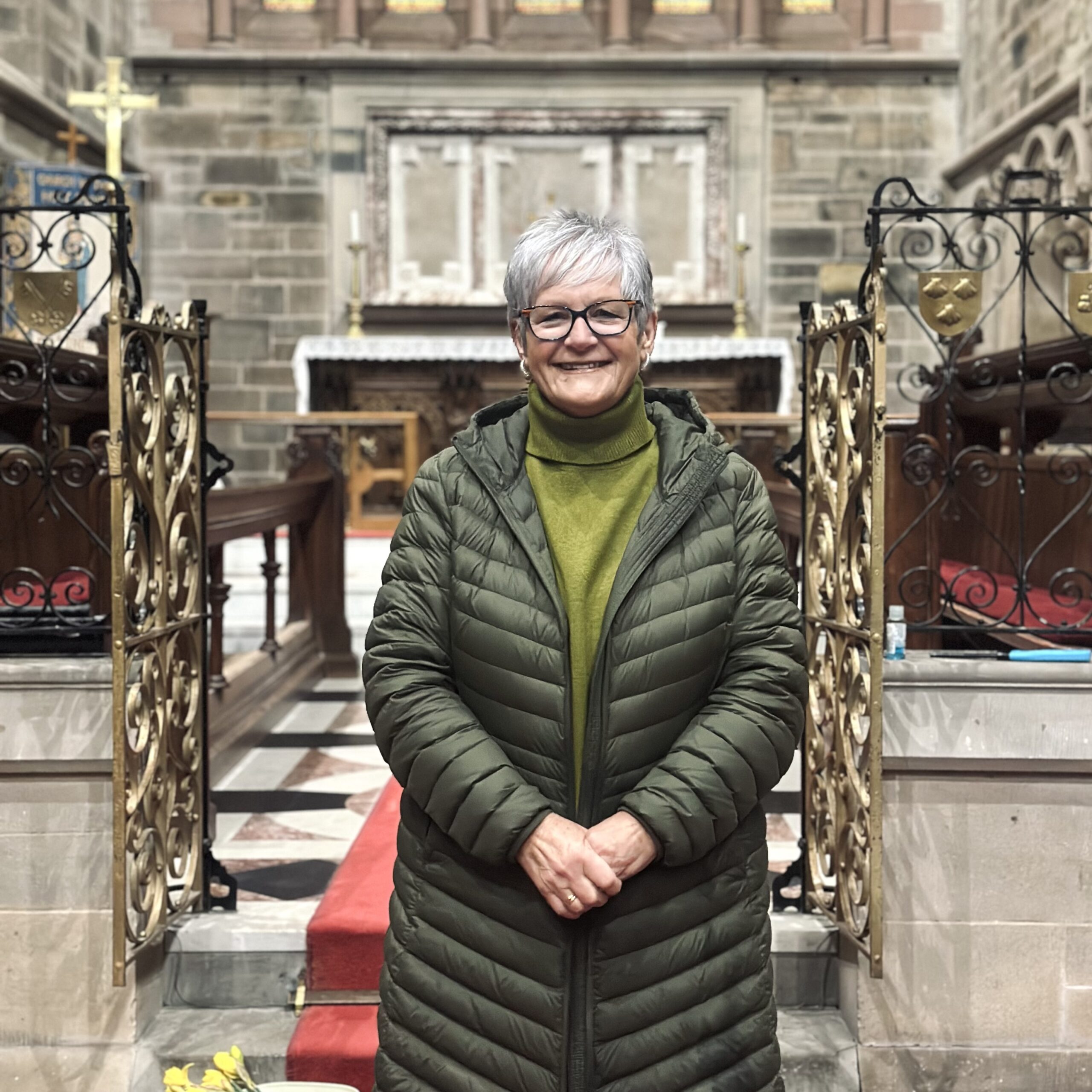An older woman with short grey hair and glasses, wearing a green jacket and polo neck, stands smiling in the centre aisle of a church with ornate stone walls and an altar in the background.