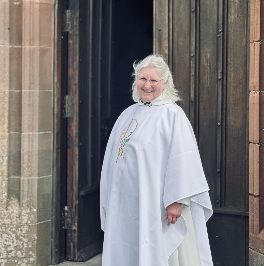 An older person with grey hair and glasses, dressed in a white clerical robe, stands and smiles in front of a large wooden door and stone building.