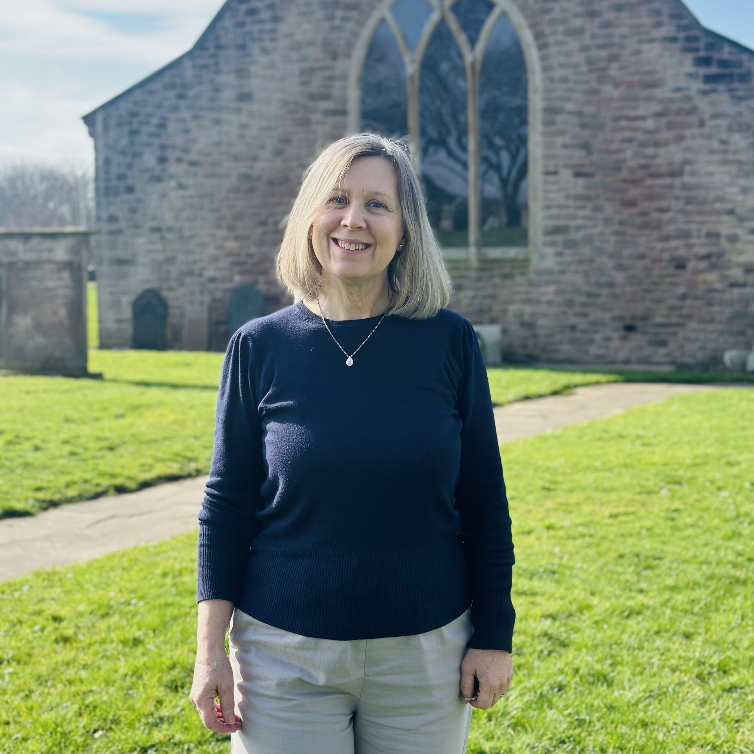 A woman with blonde hair, wearing a navy jumper and light trousers, stands smiling on green grass in front of an old stone building with arched windows on a sunny day.
